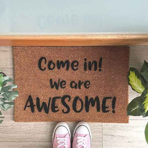 Doormat that says Come in, We are awesome. Nearby are pink shoes and green plants.