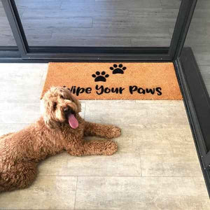 Doormat that says Wipe your Paws underneath some paw prints. There is a dog sitting next to the doormat.